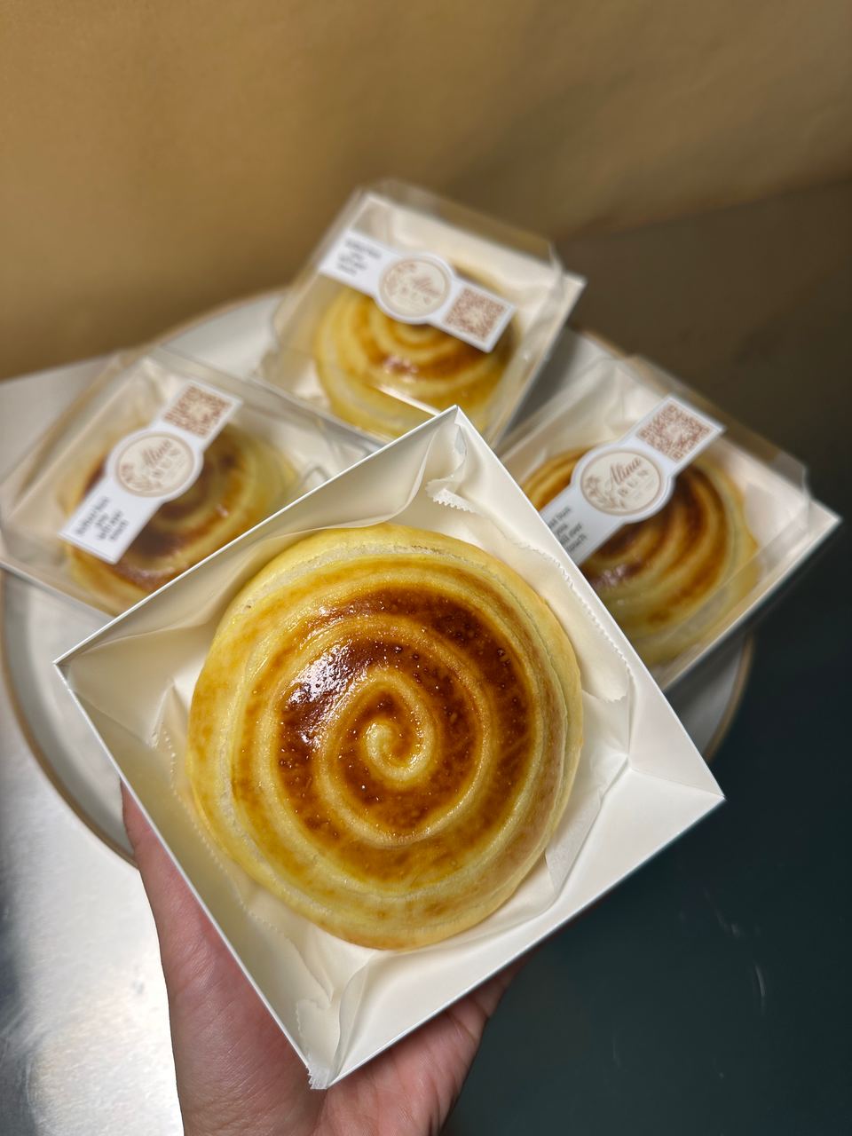A hand holding a cream bun in its clear box with the Alina Bun label