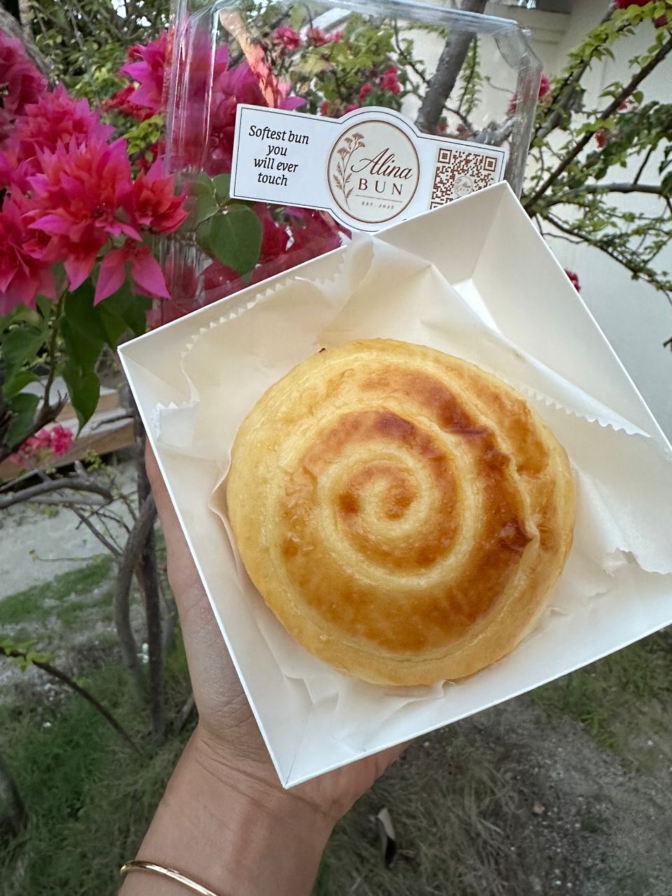 A cream bun in its box, in front of pink bougainvillea