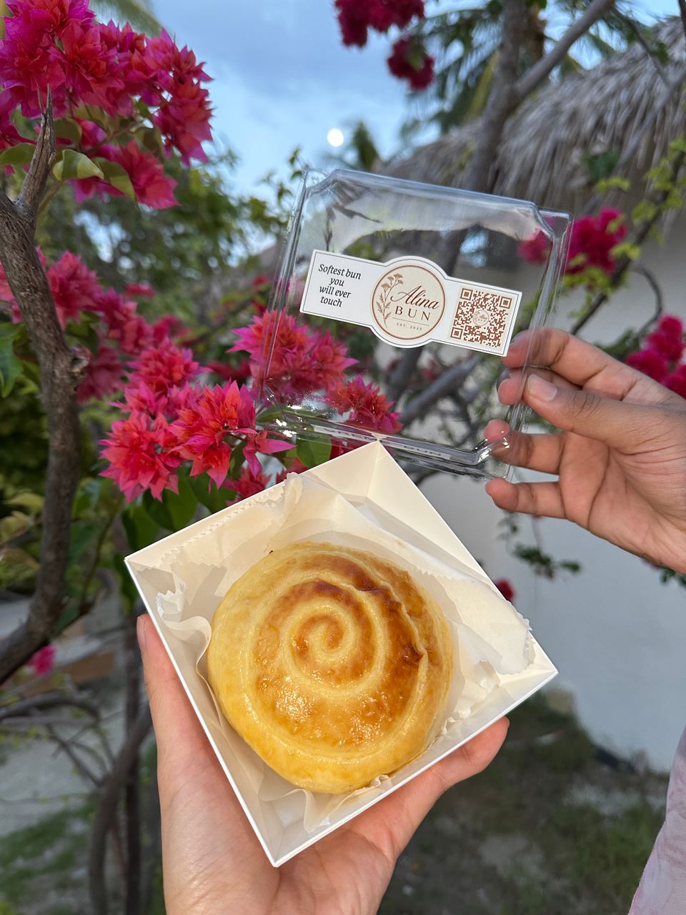 A boxed cream bun with the Alina Bun label among bougainvillea