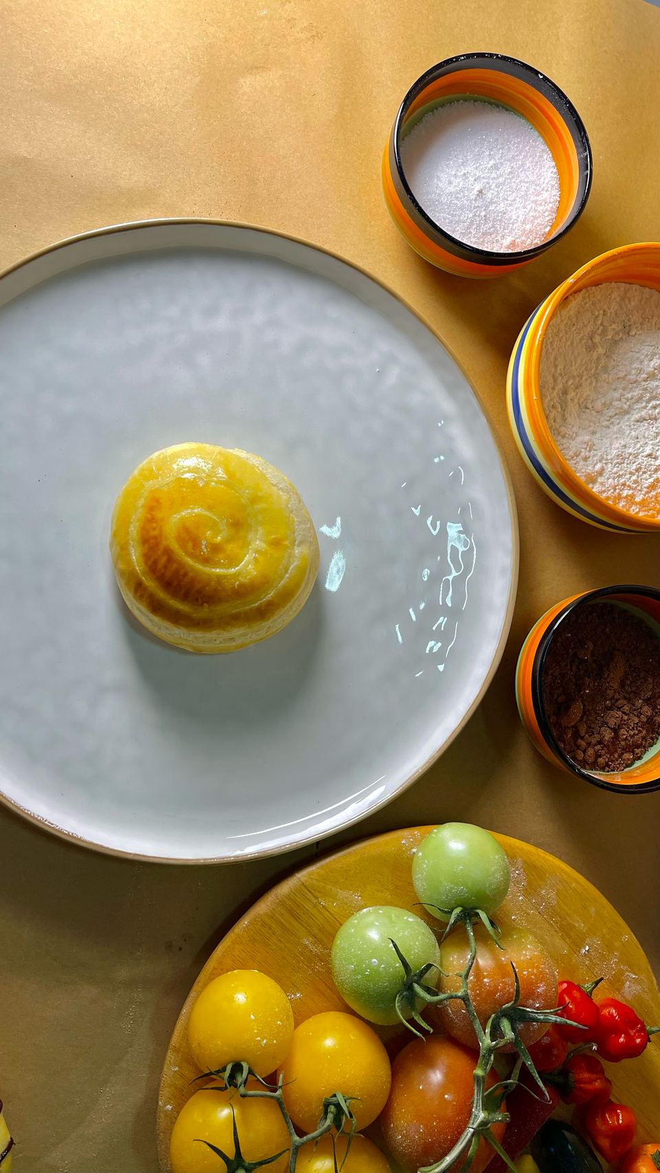 A cream bun with its golden swirl on a white plate, next to flour, sugar and tomatoes
