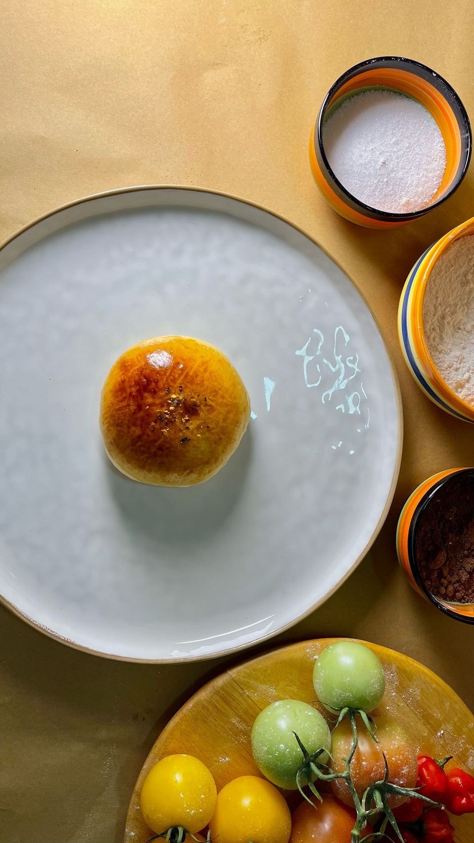 A tuna bun alone on a white plate, with bowls of flour and sugar and fresh tomatoes